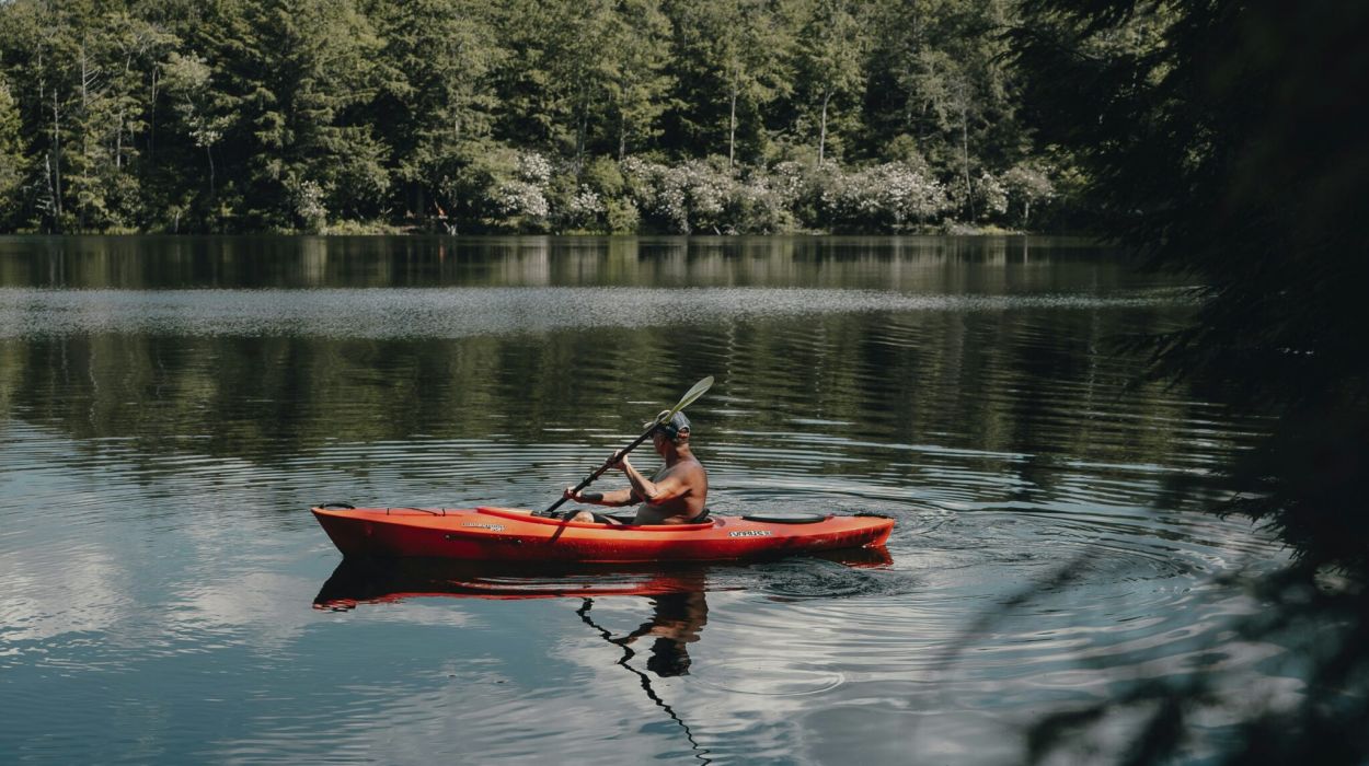 The Lodges of Huntersville A man paddles a red canoe across a serene lake surrounded by trees and mountains in the background.