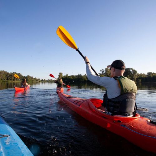 The Lodge of Huntersville A man and woman kayaking together on a serene lake, surrounded by trees and clear blue skies.