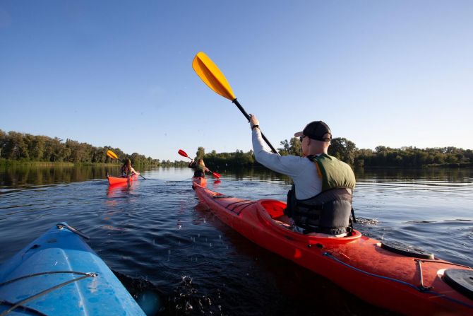 The Lodge of Huntersville A man and woman kayaking together on a serene lake, surrounded by trees and clear blue skies.